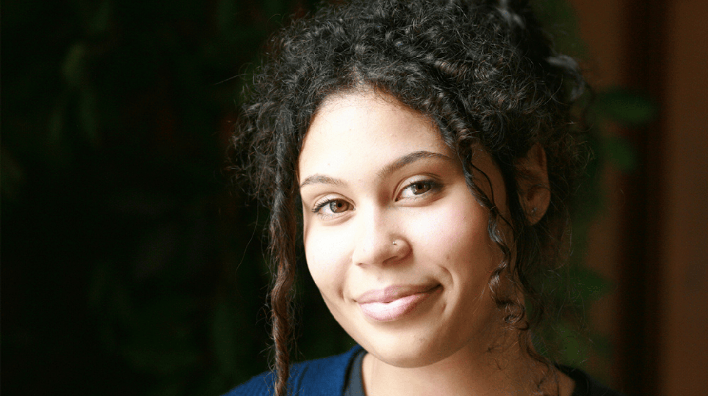 Portrait of a current Gannon Scholar with curly dark hair styled up, smiling softly, wearing a blue top, with greenery blurred in the background.